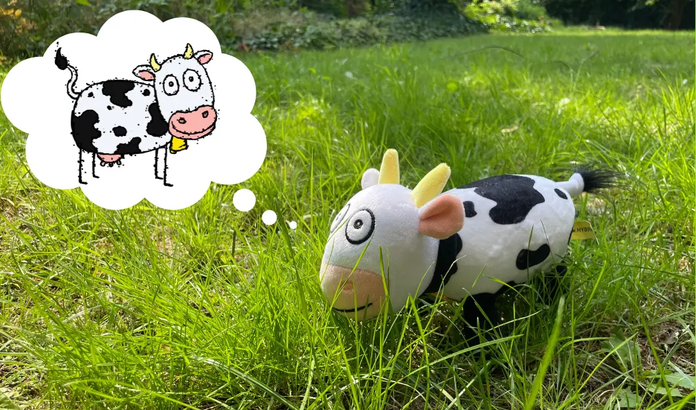 Eine Plüschkuh liegt im grünen Gras in einem sonnigen Garten. In einer Gedankenblase ist eine gezeichnete Kuh mit Glocke – das Vorbild für das Stofftier.